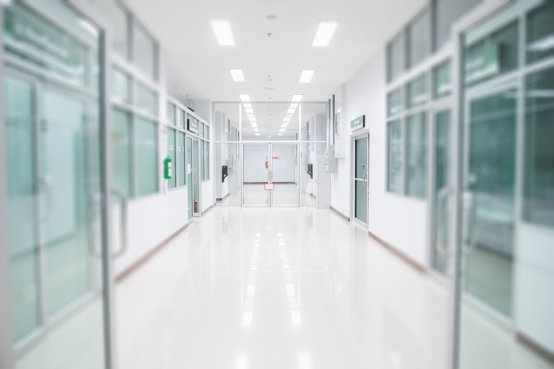 Bright, white hospital hallway with glass-walled rooms on each side.