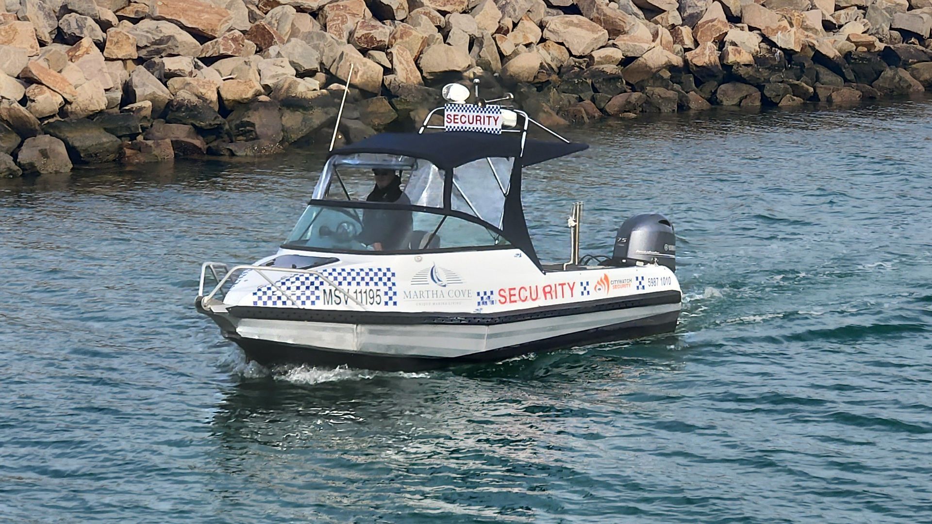 A silver boat with blue and white markings on the water, in front of a rocky shoreline.