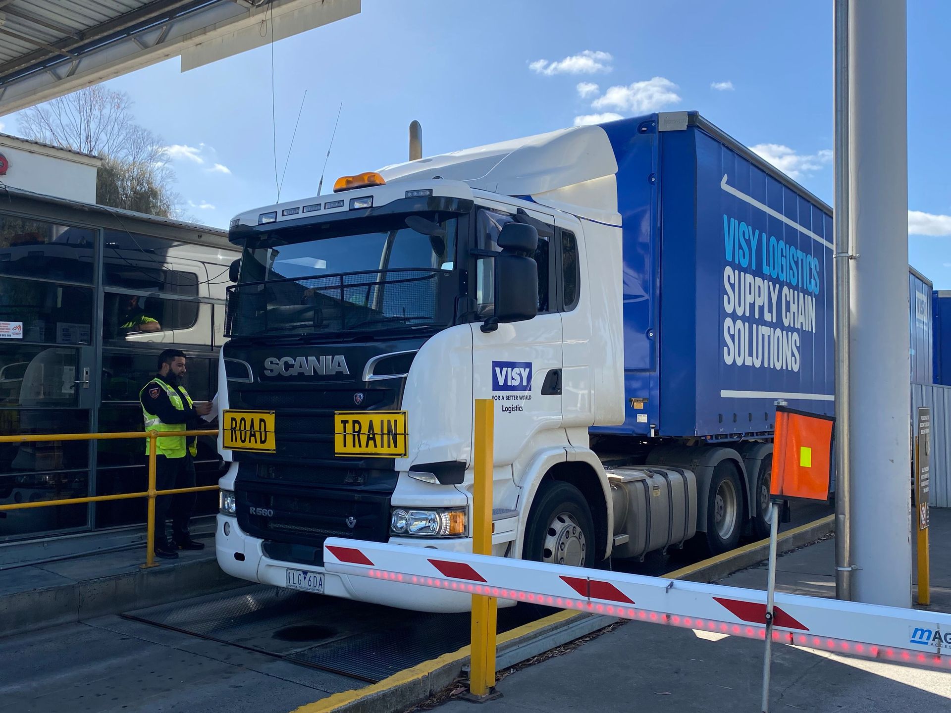 A large white and blue truck is stopped at a checkpoint. A person in a reflective vest stands beside it.
