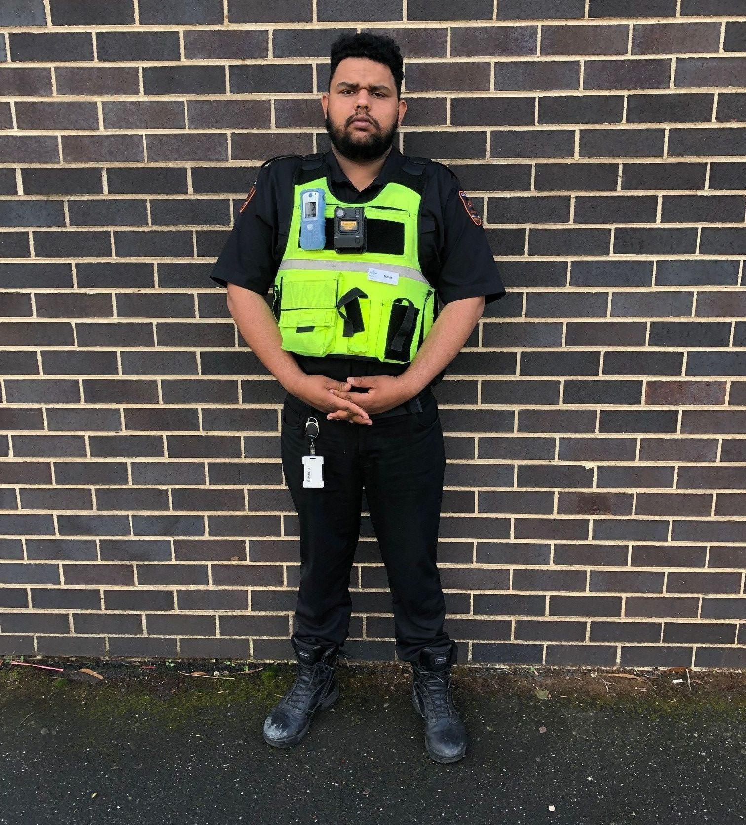 Man in black uniform and neon vest stands in front of a brick wall with hands clasped.