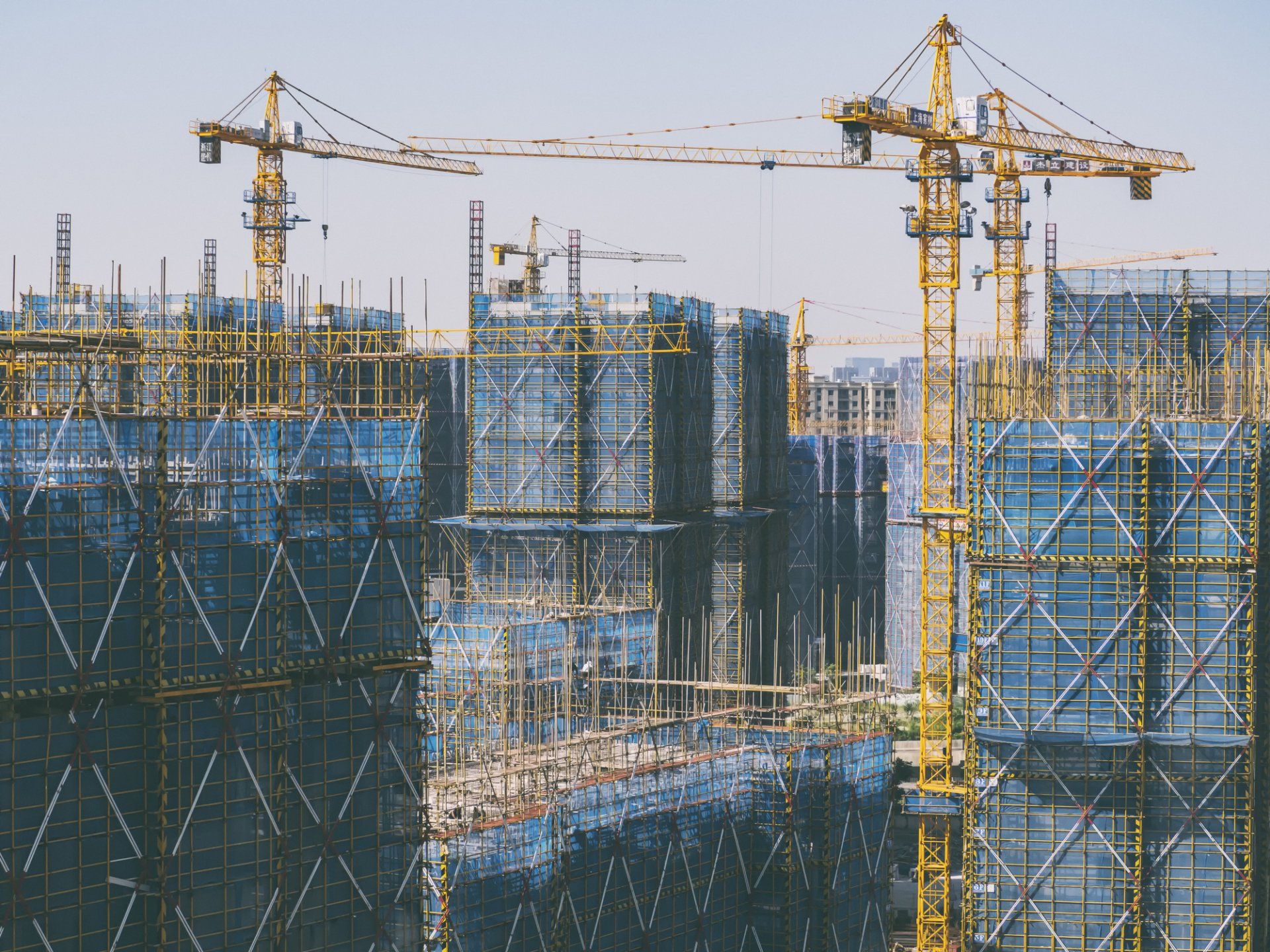 Construction site with yellow cranes and scaffolding, buildings wrapped in blue netting.