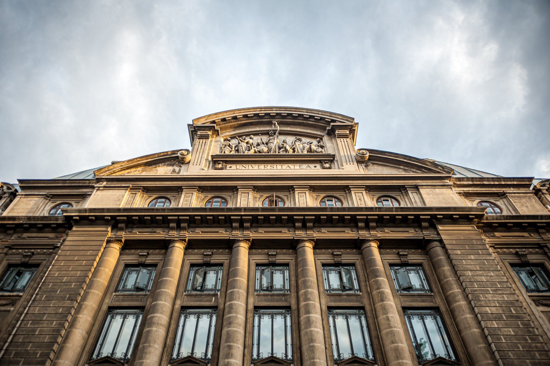 Low-angle shot of a tan building with arched windows, columns, and ornate sculptures against a cloudy sky.