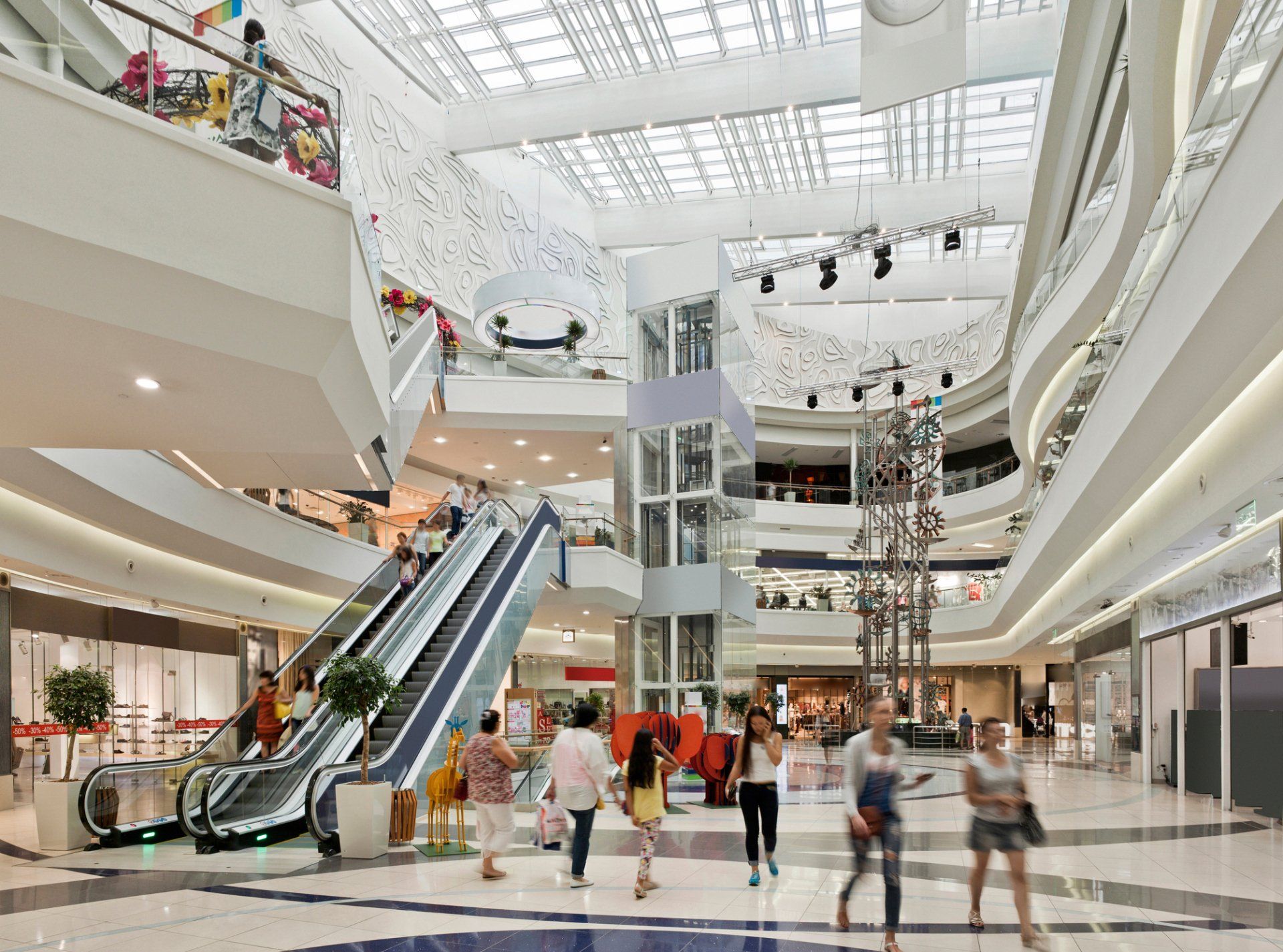 Interior view of a brightly lit shopping mall with escalators, elevators, and people walking around.