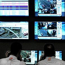 Two people monitor screens showing surveillance footage and data; a control room setting.