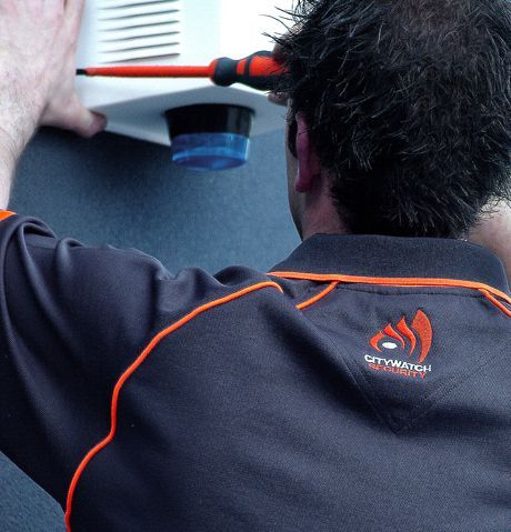 Man in grey uniform installing equipment, using a screwdriver. Logo on back says